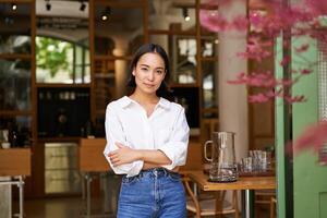 Portrait of young confident asian businesswoman, cross arms on chest, looking confident, standing near cafe entrance photo