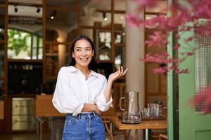 Stylish young asian woman standing in front of cafe entrance, inviting people, businesswoman owner of restaurant, looking confident and happy photo