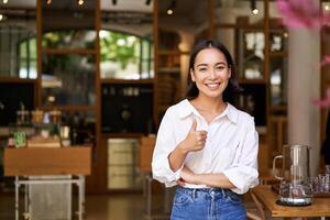 confidente asiático mujer de negocios, demostración pulgares arriba, en pie cerca Entrada de su café o restaurante, recomendando sitio foto