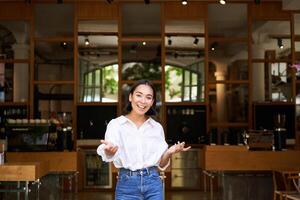 Enthusiastic young businesswoman, asian girl showing her business, raising hands up and smiling, standing in front of restaurant photo