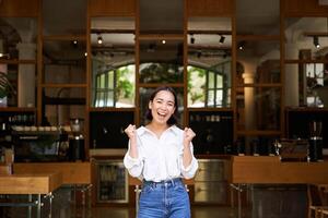 Happy enthusiastic asian girl, business owner triumphing, opening her own restaurant, standing in front of empty cafe photo