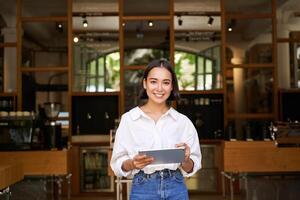 Portrait of asian woman, manager standing with tablet in front of cafe entrance, welcomes guests photo