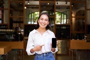 Happy asian woman, cafe manager standing in front of restaurant, pointing finger at smartphone app, showing mobile screen, recommending application photo