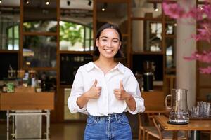 Happy asian woman shows thumbs up, approves, likes smth good, stands near cafe entrance and give her recommendation photo