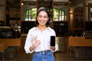 entusiasta joven asiático chica, camarera desde café demostración móvil teléfono solicitud, señalando a teléfono inteligente pantalla foto