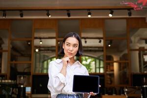 joven asiático mujer mira considerado, soportes en vacío cafetería, muestra su digital tableta pantalla foto