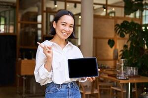 sonriente asiático mujer de negocios, demostrando, demostración tableta pantalla y señalando a bandera, publicidad foto