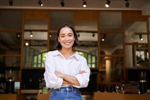 Portrait of confident young asian woman, business owner, cross arms on chest, looking pleased, standing in front of cafe photo