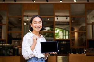 sonriente asiático mujer demostración digital tableta pantalla, café propietario demostración algo, en pie en frente de café Entrada foto