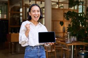 sonriente asiático mujer muestra tableta pantalla y mira complacido, soportes en café foto