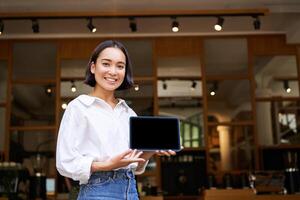 Smiling asian woman showing digital tablet screen, cafe owner showing smth, standing in front of cafe entrance photo