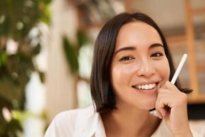 Close up portrait of stylish young brunette woman, sitting with graphic pen and smiling, relaxing in cafe, writing something, making notes photo