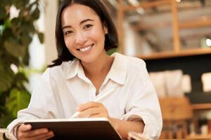 Portrait of asian girl drawing on digital tablet, scatching and doodling with graphic pen, sitting in cafe and smiling photo