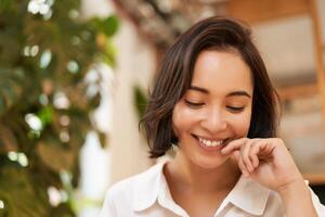 Close up portrait of beautiful, romantic young asian woman, smiling and looking happy, sitting in cozy cafe photo