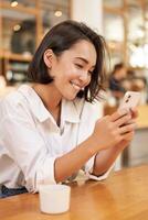 Vertical shot of brunette asian girl sitting in cafe, using smartphone app, chatting on mobile phone and smiling photo