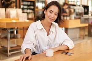 Portrait of confident asian woman, sitting in cafe, smartphone and coffee on table. Businesswoman smiling with confidence photo