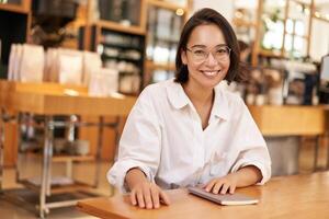 Young stylish asian woman, business owner in glasses, sitting in cafe with notebook, smiling at camera photo