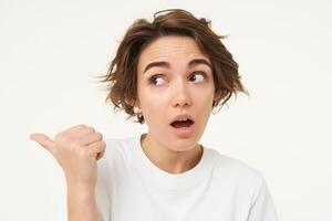 Image of girl with surprised face, points finger left, looks shocked, poses over white studio background photo