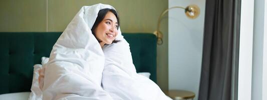 acogedor mañanas. contento asiático niña siente calentar en su cama, cubre sí misma con acogedor funda Nordica en su dormitorio, un cómodo permanecer en hotel habitación foto