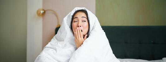 Close up of funny sleepy girl, asian woman yawns after waking up early morning, covers herself with blanket duvet, holds hand near opened mouth photo