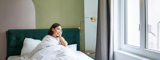 Happy korean woman wakes up in her hotel room, lying in bed under cozy warm blankets, white sheets, enjoying weekend morning photo