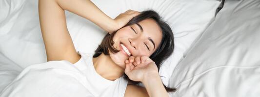 cerca arriba de contento asiático niña acostado en cama en su almohada, sonriente y laminación terminado como sensación energético en mañana, despertar arriba temprano foto