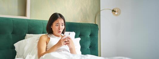 Portrait of shocked korean woman looking at her mobile phone, lying in bed, reading bad shocking news on smartphone app photo