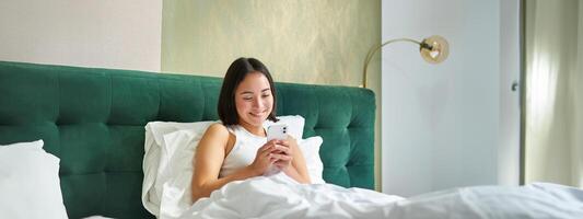 Cute korean girl in bed, holding smartphone, feeling happy and pleased, spending morning in bed, enjoying surfing net on mobile phone photo