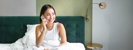 Smiling korean girl in bed, talks on mobile phone, making a phone call, lazy morning as asian woman orders delivery via smartphone photo