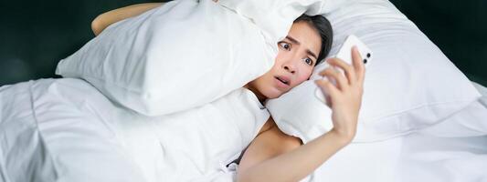 retrato de asiático mujer despertar arriba en cama, mirando conmocionado a móvil teléfono, darse cuenta ella dormido foto