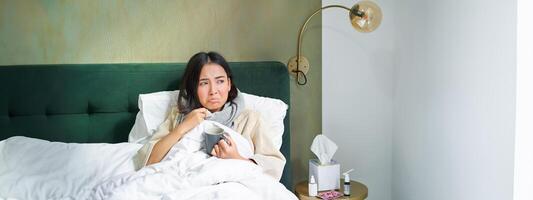 Covid-19, flu and vaccination concept. Korean girl lying in bed with cold, catching influenza, drinking hot tea and using prescribed medication photo