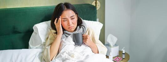 retrato de asiático mujer con dolor de cabeza, atrapando columna, quedarse en enfermo salir a hogar, acostado en cama, Bebiendo caliente té, teniendo gripe foto