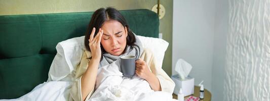 gripe, enfermedad y gente. joven mujer sensación enfermo, teniendo fiebre y atrapando columna, acostado en cama con caliente té, tomando drogas desde influenza foto