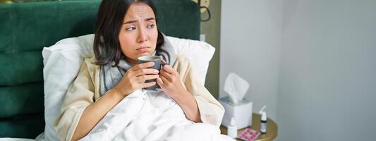 COVID-19, gripe y vacunación concepto. coreano niña acostado en cama con frío, atrapando influenza, Bebiendo caliente té y utilizando prescrito medicación foto