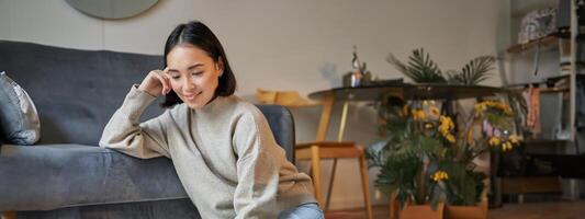 Vertical shot of young woman in cozy home working on laptop, using smartphone and drinking coffee, sitting on floor near sofa photo