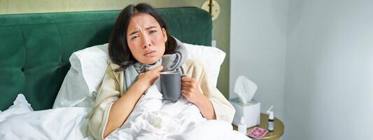 retrato de acostado asiático niña en cama, sensación enfermo, atrapando frío y quedarse a hogar, mirando insalubre, Bebiendo caliente té foto