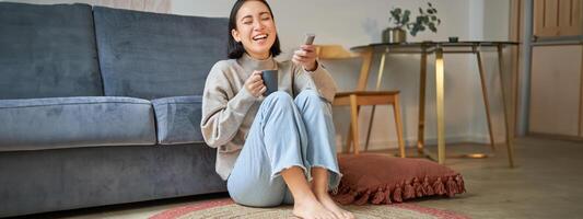 Portrait of young woman with remote, watching tv, switching chanels on television, sitting on floor near sofa and relaxing photo