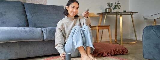 Portrait of beautiful asian girl sitting at her home and watching tv, holding remote, smiling and laughing, feeling comfort and warmth at her apartment photo