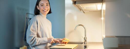 Portrait of smiling young asian woman standing on kitchen and making a sandwitch, cooking for herself photo