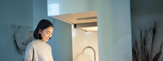 Vertical shot of young asian woman cooking dinner, making herself sandwitch, smiling while standing on the kitchen photo