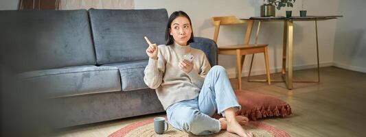 Portrait of woman sitting on floor with smartphone, looking thoughtful and pointing finger at banner, promo advertisement on top right corner photo