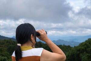 Young woman with binoculars on the mountain on a sunny day. Woman using binoculars when going hiking. Hiking woman uses binoculars to travel and has a happy smile. photo