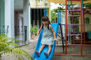 contento niña jugando en el deslizar. contento pequeño asiático niña corredizo y jugando a al aire libre patio de recreo en parque en verano vacaciones. sano actividad. foto