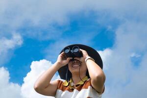 Young woman with binoculars on the mountain on a sunny day. Woman using binoculars when going hiking. Hiking woman uses binoculars to travel and has a happy smile. photo