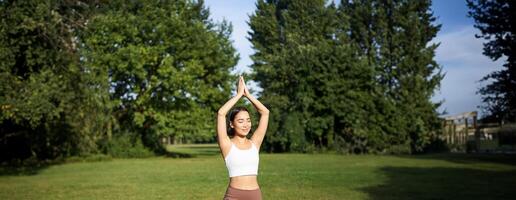 vertical Disparo de asiático mujer en pie en asanas, haciendo yoga ejercicios en Fresco aire en parque, vistiendo polainas, en pie en caucho estera foto