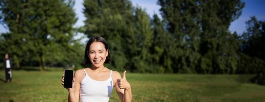 Vertical shot of asian woman shows thumbs up, recommending yoga training online, meditation app, doing exercises on fresh air in park, showing smartphone screen photo