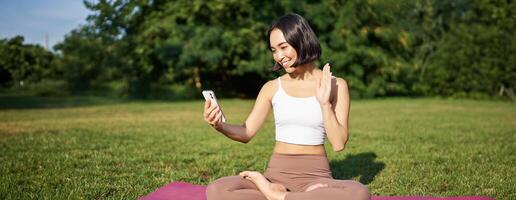 Portrait of asian girl say hello on smartphone, wave at mobile phone while doing meditation, online yoga classes on fresh air in park photo