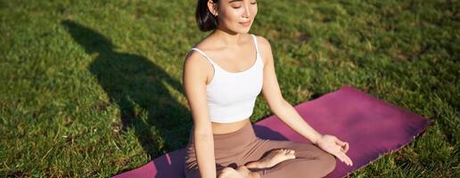 Portrait of smiling asian woman meditating, doing yoga on fresh air, relaxing on rubber mat, exercising in park, breathing air, being calm photo