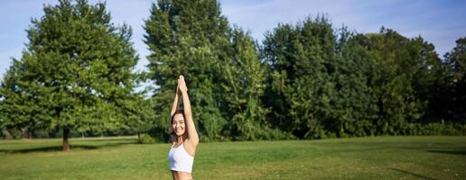 Vertical shot of smiling korean woman doing tree yoga asana, stretching on rubber mat in park, exercising photo