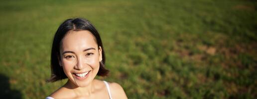 Vertical shot of young asian woman doing yoga, practice mindfulness, smiling and looking relaxed, sitting on rubber mat in park photo